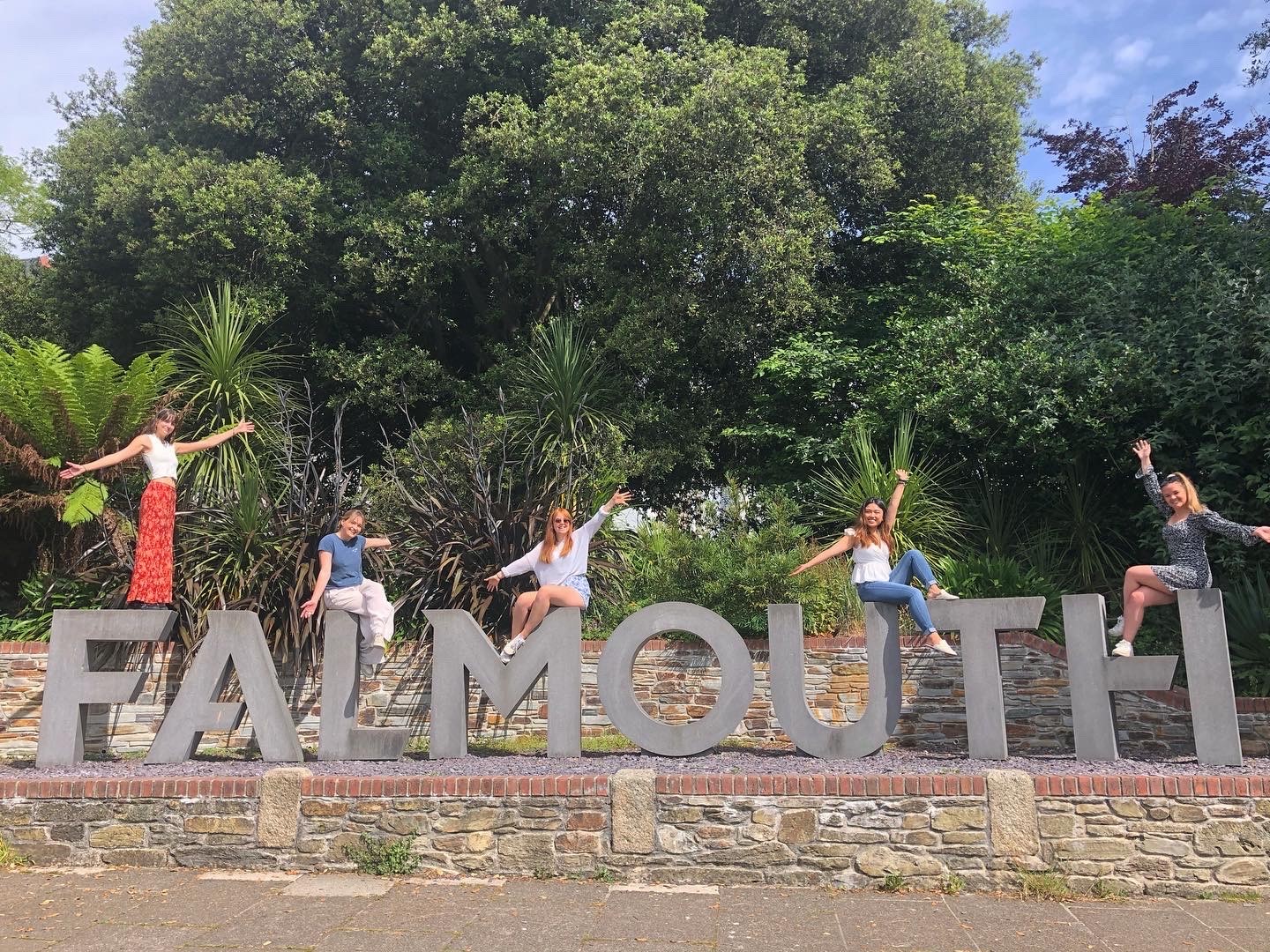 Students sit on the Falmouth sign opposite Woodlane campus - large, grey concrete letters that spell out 'Falmouth'